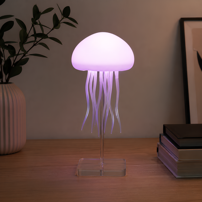 A jellyfish-shaped table lamp with a glowing purple-white dome and translucent tentacles on a clear acrylic stand, placed on a wooden tabletop with a vase and books in the background.